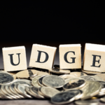 The word 'Budget' spelled out using blocks, resting on a bed of gold coins.